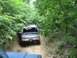 1989 GMC Jimmy - A dark SUV navigates a narrow dirt path surrounded by dense trees, with another vehicle partially visible ahead.