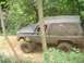 1989 GMC Jimmy - A muddy SUV with a camper shell navigates a dirt path surrounded by dense foliage.