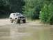 1989 GMC Jimmy - A 1989 GMC Jimmy drives through shallow water on a dirt path surrounded by trees.