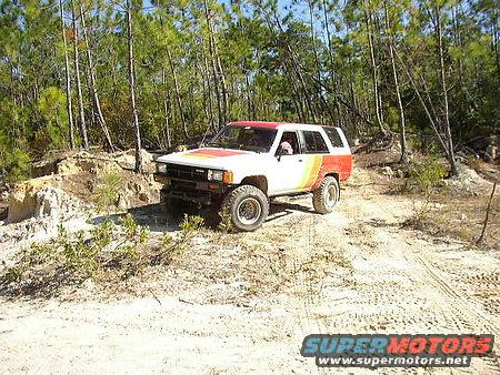 White and red 1984 Toyota 4Runner off-roading on a dirt path with trees in the background