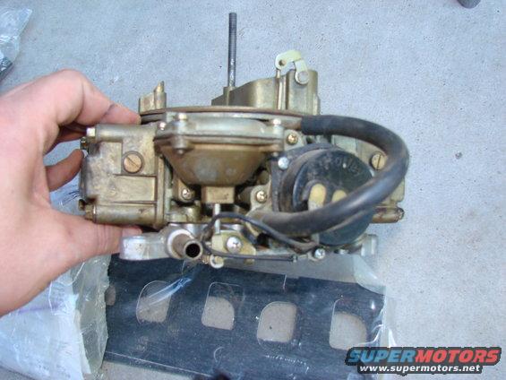 Hand holding a Holley carburetor. Black hose attached to the side
