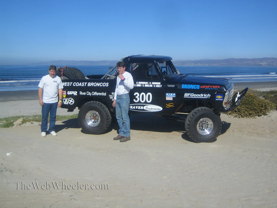 Black Ford Bronco with racing decals and two men standing beside it on a sandy beach
