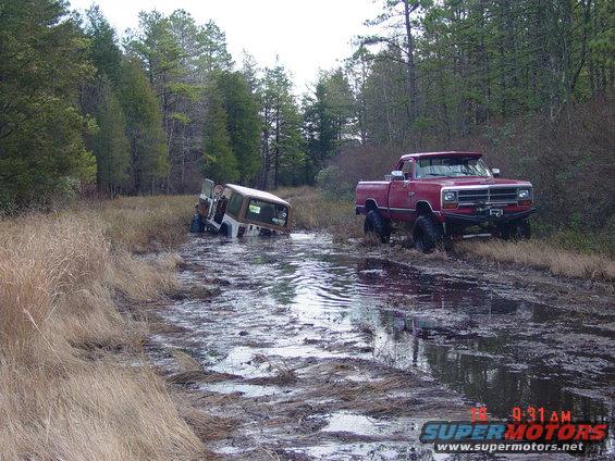 Red 1990s Dodge pickup truck with large tires in mud next to a partially submerged SUV
