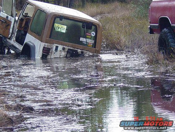 Brown Jeep Wrangler TJ rear view, partially submerged in water with a red truck visible to the right