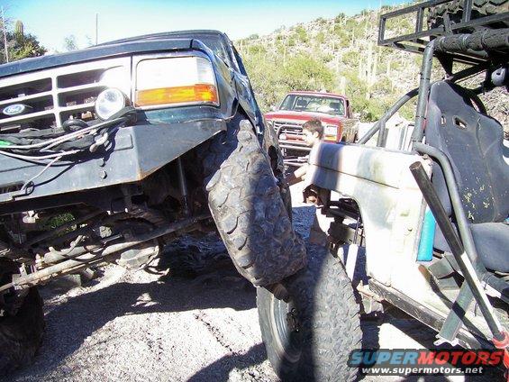 Front of a dark green Ford Bronco and a buggy-style off-road vehicle are close together. A red pickup truck is behind them