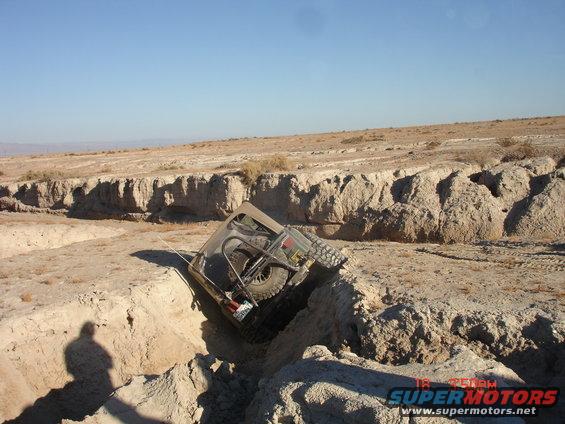 Jeep-like vehicle tilted in a dry ditch, spare tire visible on the back. Sunny day, desert landscape