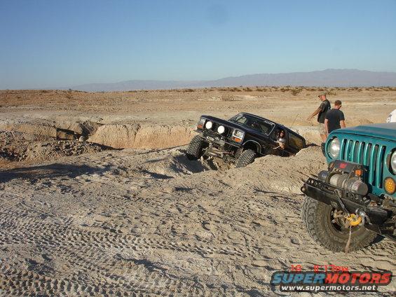 Black Ford Bronco stuck in a sandy ditch, pulled by a teal Jeep. Desert landscape