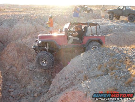 Red Jeep Wrangler climbing rocky terrain with people watching. Another Jeep and a pickup truck are visible in the background