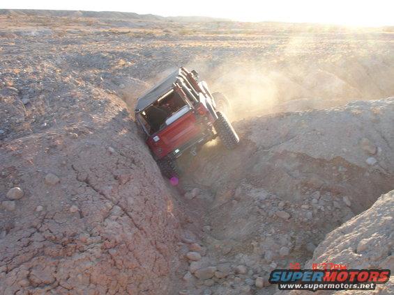 Red Jeep Wrangler going down a steep dirt ravine, kicking up dust. Sun glare in the background