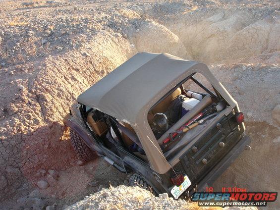 Jeep Wrangler tilted in a dirt ditch, soft top up, gear in back. License plate K95 ACE