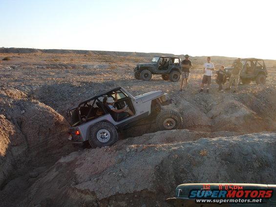 Several Jeeps navigating a rocky off-road course under a clear sky. One Jeep is on a steep incline
