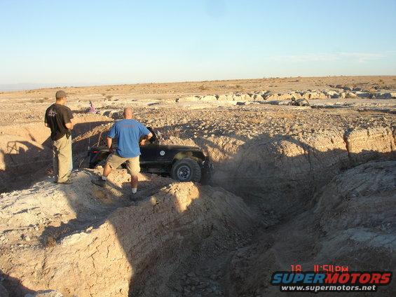Two men watch an off-road vehicle attempt to drive through a deep dirt trench in a desert landscape