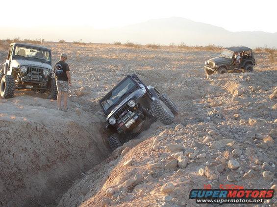 Three Jeeps navigating a rocky ditch in a desert landscape. One Jeep is stuck in the ditch