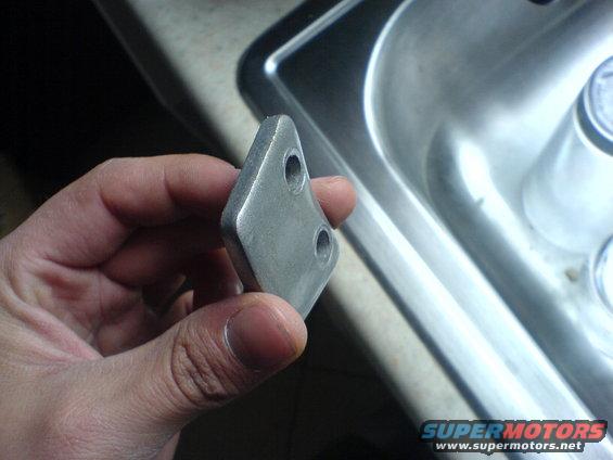 Hand holding a small, diamond-shaped metal part with two holes near a sink