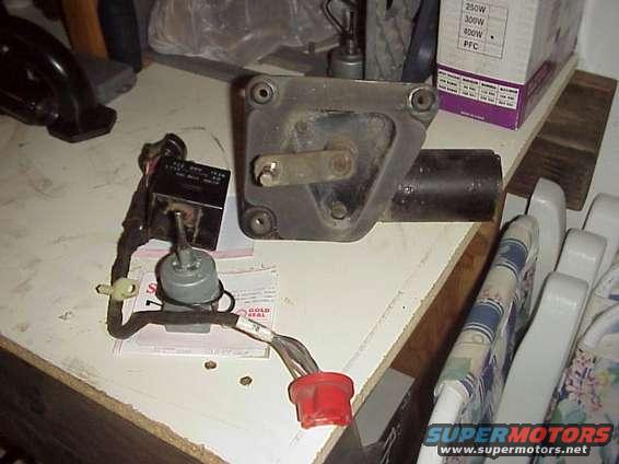 Windshield wiper motor and switch assembly on a workbench. The motor is black and metal, with a lever arm. The switch is gray with a black box and red connector