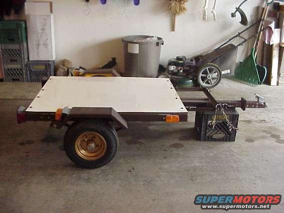 Utility trailer with white deck, brown fenders, and one wheel visible. Lawn mower and trash can in background