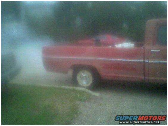 Side view of a red pickup truck with some stuff in the bed. Another vehicle is partially visible on the left, emitting smoke