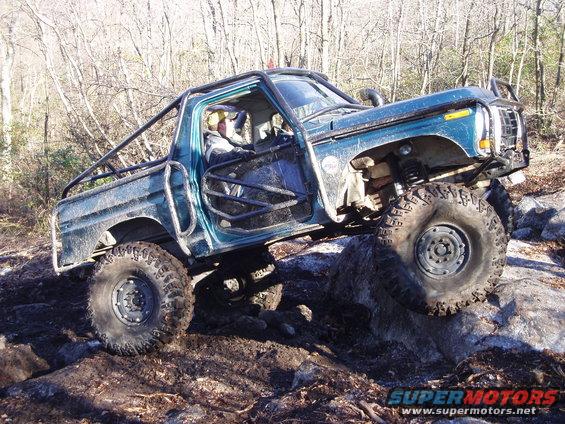 Modified pickup truck with roll cage climbing over rocks off-road. The front grille and body style suggest a late 1970s Ford F-Series