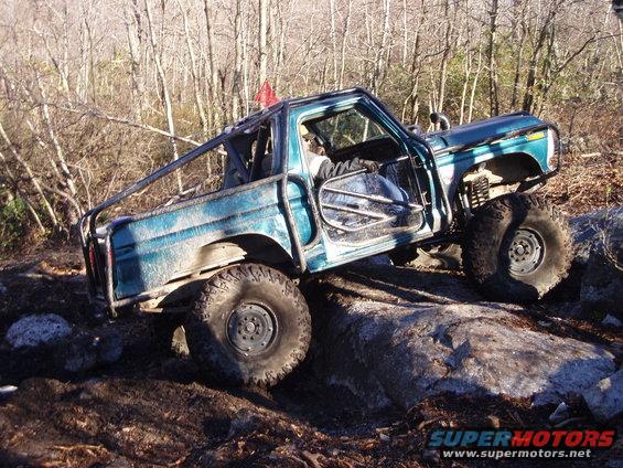 Teal pickup truck with roll cage climbing over rocks on a wooded trail