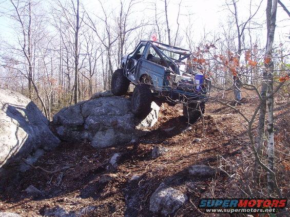 Modified off-road truck with a roll cage climbing over large rocks in a wooded area