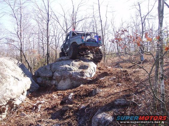 Teal off-road truck with roll cage climbing over large rocks on a wooded trail