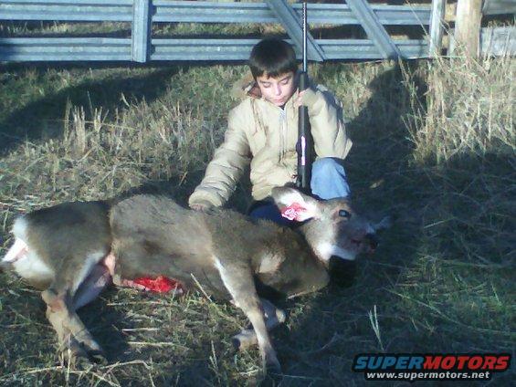Deer carcass on grassy ground with a boy holding a rifle next to it