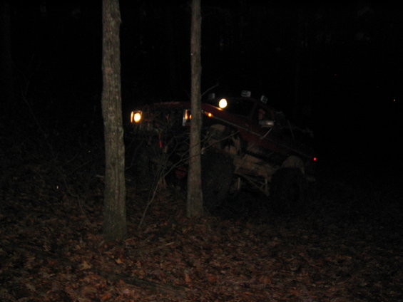 Red off-road vehicle with auxiliary lights on, driving through trees at night