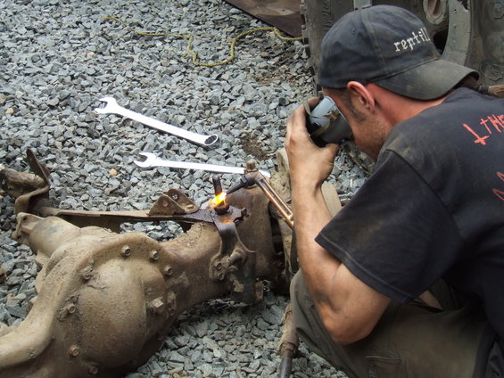 Guy using a torch to weld a bracket onto a dirty vehicle axle. Two wrenches are on the ground nearby