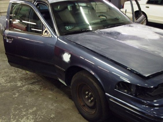 Dark blue sedan, driver's side doors open, hood up, primer on fender, missing headlight