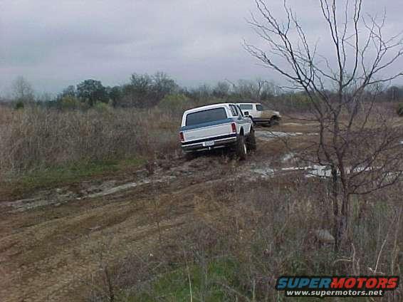 1990 Ford Bronco and another SUV driving through mud. The Bronco is white with a blue stripe