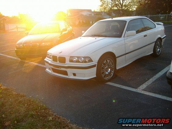 White BMW E36 coupe parked in a lot, sun glare on front. Dark BMW sedan behind
