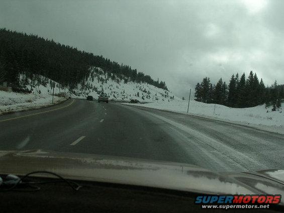 Dashboard view of a highway with snow-covered trees and two cars driving on a wet road