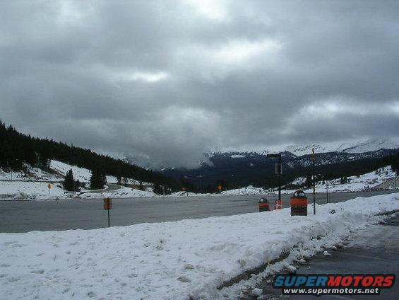 snowy mountain landscape with a parking lot and overcast sky. Supermotors.net logo in corner
