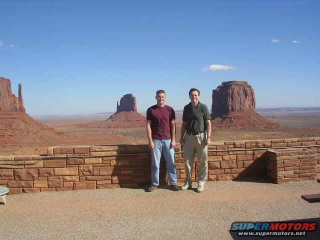 Two men stand before Monument Valley rock formations under a clear blue sky