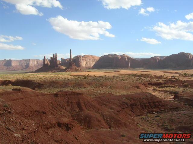 Monument Valley with rock formations under a blue sky with clouds. Supermotors.net logo in corner