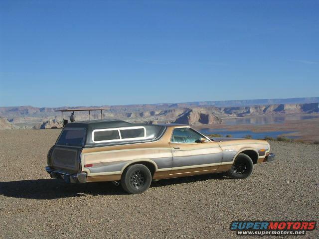1975 Ford Ranchero with a camper shell, tan and grey, parked on a gravelly overlook with a lake and canyon in the background