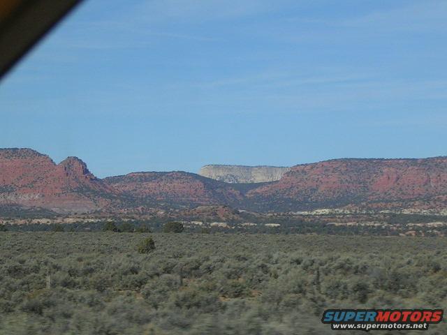 Desert landscape with red rock formations and scrub brush under a blue sky. Watermark reads SUPERMOTORS