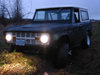 Dark 1968 Ford Bronco with headlights on, parked in a field