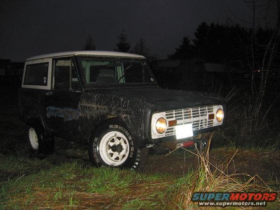 Black 1968 Ford Bronco with white roof and white wheels, muddy, parked in grass at night