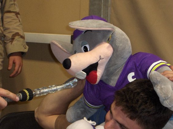 Chuck E. Cheese mascot costume with a purple shirt with a white C, held by a person, with another person's hand holding a toy