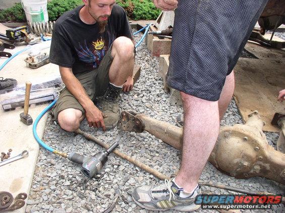 Two men are working on vehicle axles and suspension parts on a gravel surface