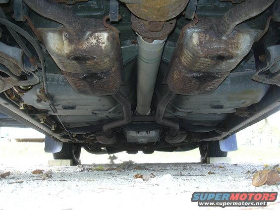 Underside of a car showing dual exhaust mufflers, driveshaft, and fuel tank. The vehicle is raised on blocks