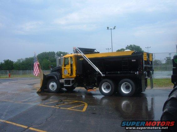 Yellow and black snow plow truck with an American flag on the plow, parked on wet asphalt