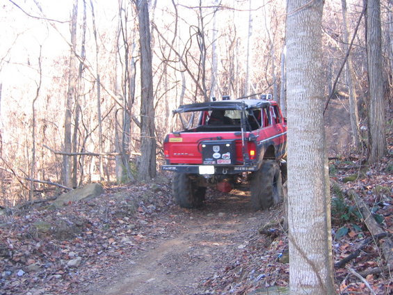 Red off-road SUV with large tires and roll cage, driving on a dirt path through trees
