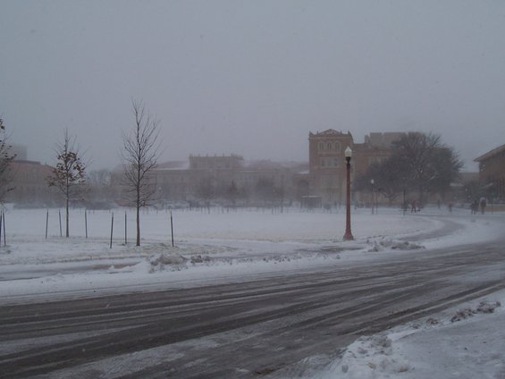 Snowy street with tire tracks, bare trees, and buildings in the background under a hazy sky