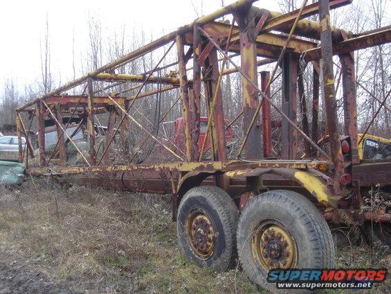 Rusty logging trailer with dual rear wheels, parked in a junkyard with other vehicles visible