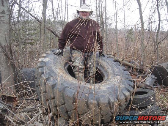 Man in a hat and brown jacket stands in a massive tire in a wooded junkyard