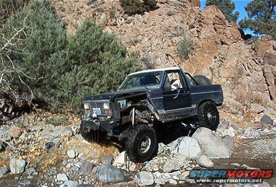 Black off-road SUV with a white top navigating rocky terrain. Front end is damaged