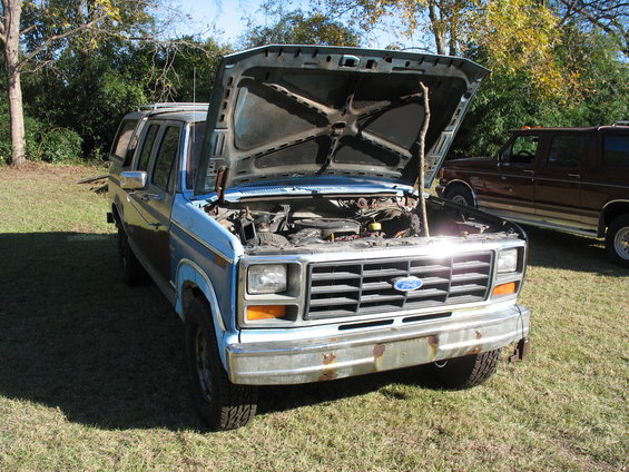 Front view of a light blue Ford truck with hood open, showing the engine. Another brown Ford truck is parked behind it