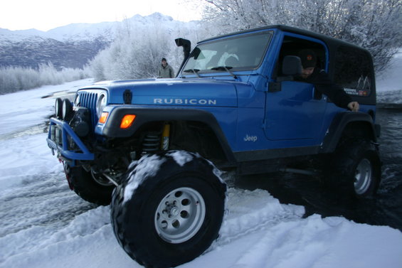 Blue Jeep Wrangler with Rubicon badging driving through snow and water, mountains in background